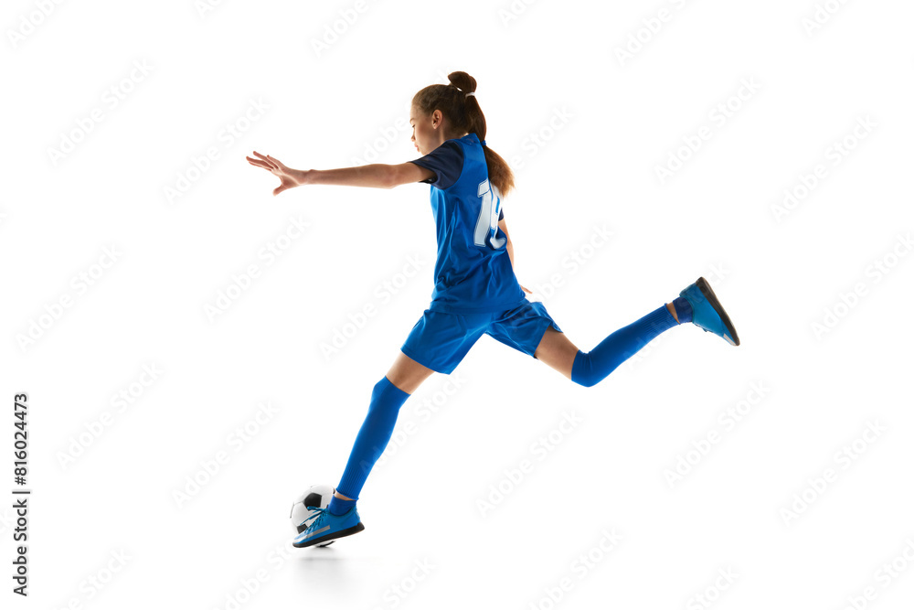 Athletic teen girl in blue uniform in motion, practicing football, kicking ball isolated on white studio background. Concept of sport, active and healthy lifestyle, childhood, school, hobby