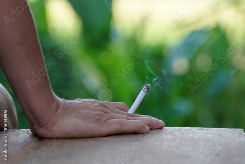 An Asian man's hand holds a lit cigarette and rests it on the wooden floor as he sits alone smoking in the smoking corner of his house.