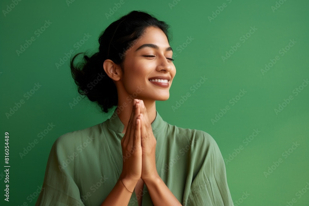 Portrait of a blissful indian woman in her 20s joining palms in a gesture of gratitude in front of soft green background