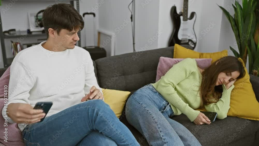A man quietly reaches for a smartphone from a sleeping woman's hand in a cozy living room, suggesting secrecy in a relationship. A man quietly reaches for a smartphone from a sleeping woman's hand in a cozy living room, suggesting secrecy in a relationship.