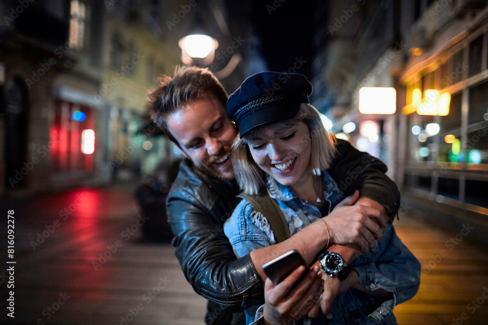Couple hugging and laughing while looking at smartphone on city street at night Couple hugging and laughing while looking at smartphone on city street at night