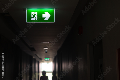 Emergency exit sign in a building corridor. A green fire escape sign is mounted on the ceiling of a dark hallway in a building near the emergency exit door.