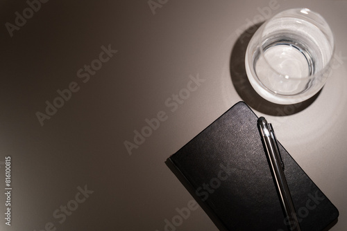Close up seen from above of small hotel table with black leather diary, pen and glass of water creating contrast of lights and shadows