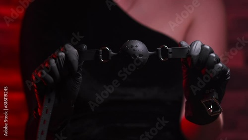 Close-up of female hands in black latex gloves and black dress holding a black BDSM gag on a red background. BDSM - outfit for sex games for adults
