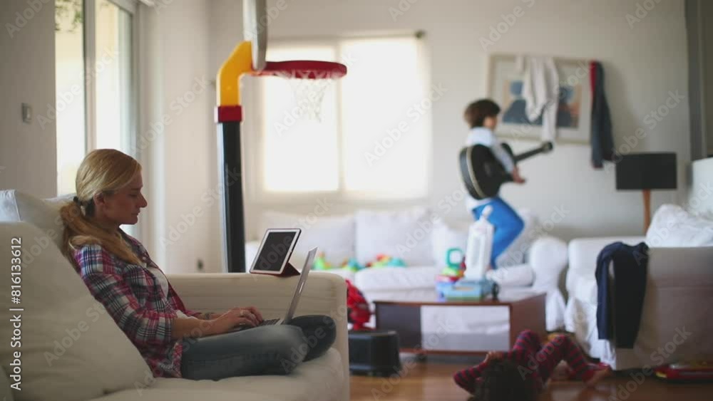 A mother works from her laptop on a couch, while her children play energetically in the living room filled with toys. A mother works from her laptop on a couch, while her children play energetically in the living room filled with toys.