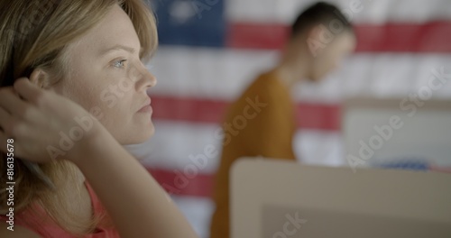 Big close up, profile, young, red-haired woman voting at booth, looking straight ahead and considering candidate choices. Asian man and US flag in background