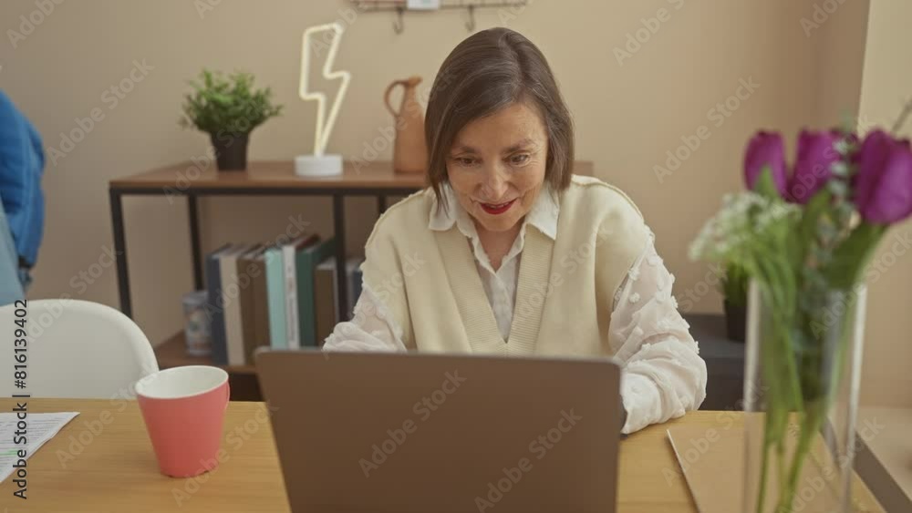 A mature woman works contentedly on her laptop in a cozy living room, surrounded by houseplants and a decorative lamp. A mature woman works contentedly on her laptop in a cozy living room, surrounded by houseplants and a decorative lamp.