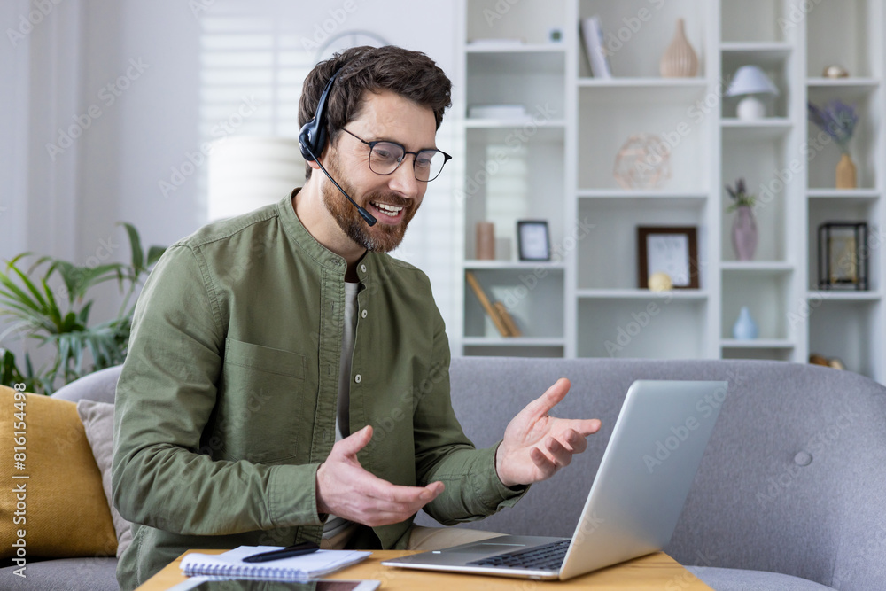 Man with a headset conducting a virtual business meeting from home, sitting on a couch with a laptop and notebook. Man with a headset conducting a virtual business meeting from home, sitting on a couch with a laptop and notebook.