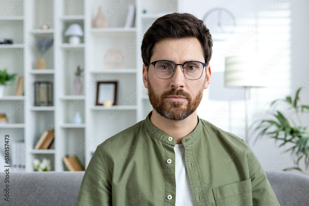 A serious man with glasses sitting in a modern home office, looking thoughtfully at the camera. Shelves with decor in the background. A serious man with glasses sitting in a modern home office, looking thoughtfully at the camera. Shelves with decor in the background.