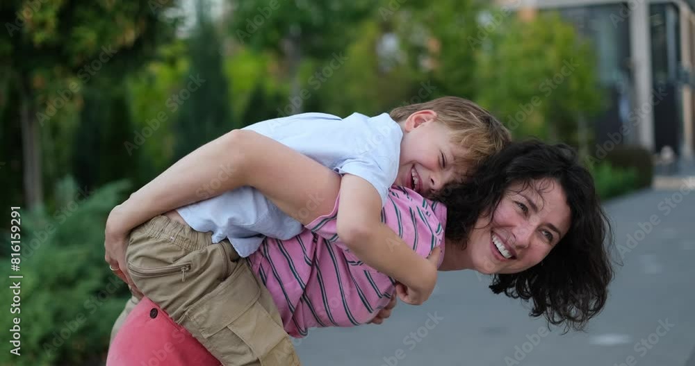 Young caucasian mother and son kid having fun together outdoors. Mother taking her child on a sholders or on the back. Bright summer day in the urban background Young caucasian mother and son kid having fun together outdoors. Mother taking her child on a sholders or on the back. Bright summer day in the urban background