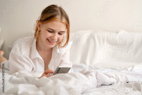 Happy cheerful smiling teenage girl laying on bed using mobile phone, scrolling social media blogs, chatting, communicating online, light airy neutral creamy bedroom interior