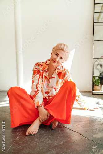 Mature blonde woman relaxing, sitting on the floor, looking at the camera with delicate smile.