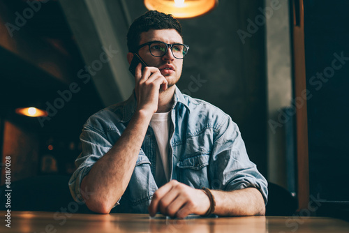 Serious concentrated man calling to operator for checking account balance using smartphone with 4g internet, pondering hipster guy in spectacles making telephone conversation with colleague indoors