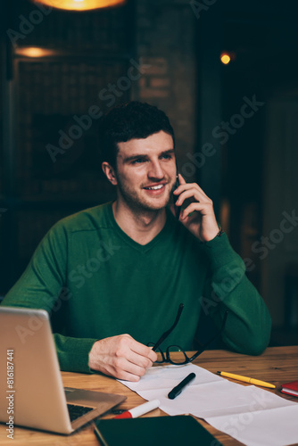 Successful male entrepreneur talking with friend in break of planning organisation and work on laptop device, happy hipster guy making positive conversation via cellular phone using application