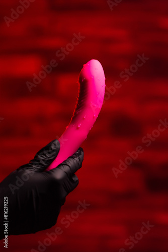 Pink stylish vibrator for masturbation in the hands of a girl in black gloves on a red background. Massager with multiple speeds. Products for sex shop, gifts for adults