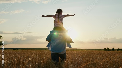 Little daughter on shoulders of her father. Girl and dad are traveling through wheat field. Child and parent play in nature. Happy family, childhood concept. Slow motion. Daughter, dad farmer walking photo