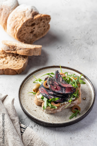 Sandwich with salami sausage, grain bread, cream cheese, olives, aragula and microgreens with ingredients on light gray textured background. Breakfast