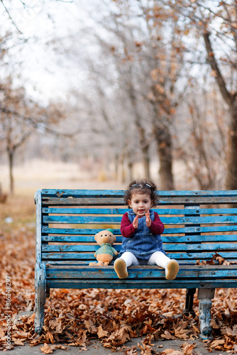 Adorable curly hair girl sitting on a peeling blue bench with a bear in autumn park and clapping hands