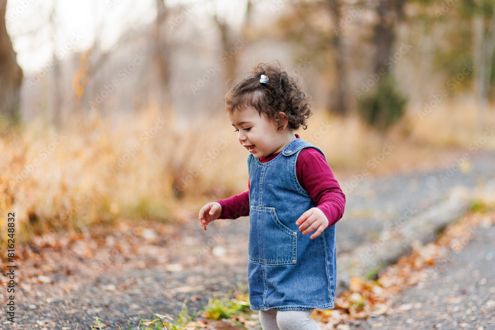 Charming little child in denim dress and maroon shirt walks on leaf-covered path in autumn