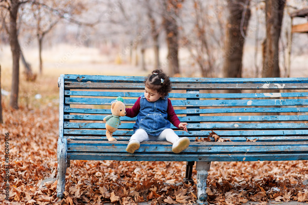 Child and teddy bear. Adorable girl sits on a worn blue bench among fallen leaves