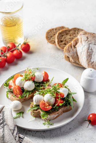 Sandwiches with tomatoes, mozzarella balls, arugula on white plate light textured background with ingredients. Italian bruschetta set, antipasti