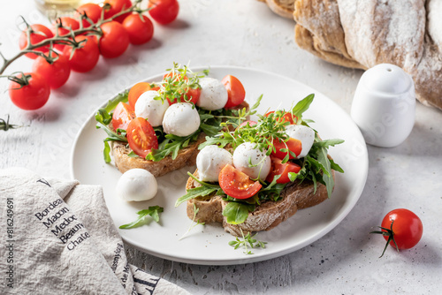 Sandwiches with tomatoes, mozzarella balls, arugula on white plate light textured background with ingredients. Italian bruschetta set, antipasti