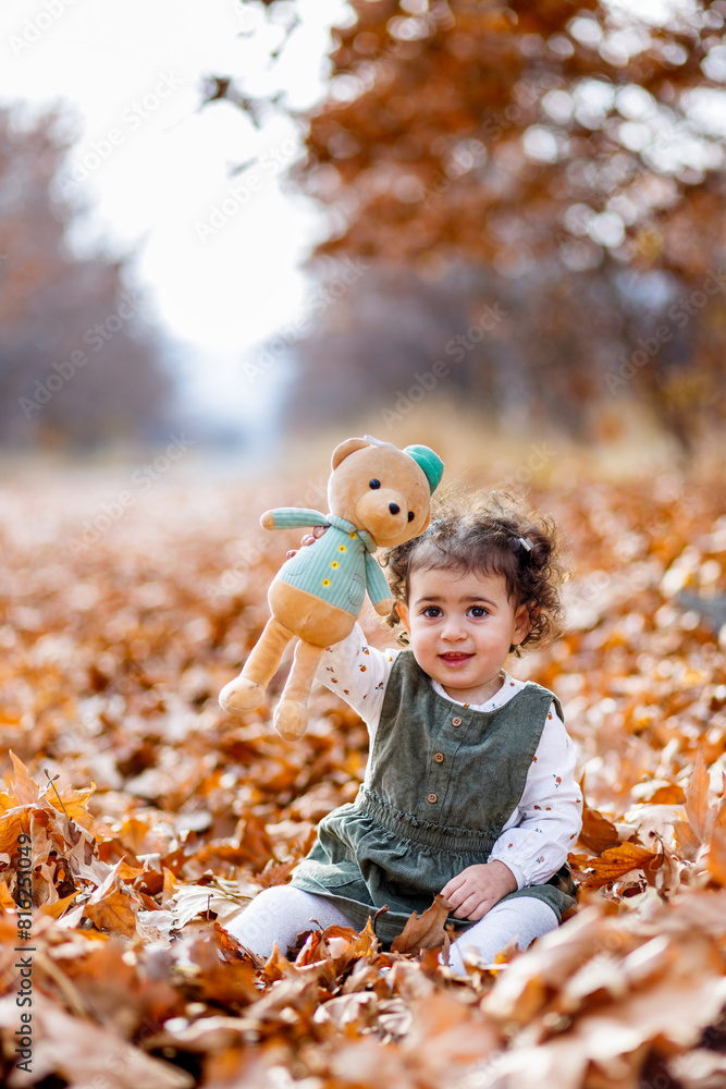 Playful autumn moments. Sweet little girl sitting among autumn leaves, showing her teddy and smiling