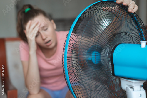 Young woman using electric fan at home in living room, sitting on couch cooling off during hot weather, suffering from heat, high temperature
