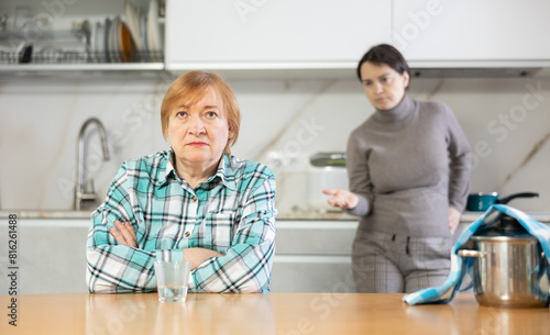 Young girl makes claims her mother emotionally.Sad mature woman with soup in pan on table listens to reproaches of daughter sitting on kitchen at home. Misunderstanding relationship photo