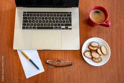 Working desk with a notebook, a red cup of coffee, some sweet cookies on a plate, a pair of glasses, and a blue and gold fountain pen, on a sheet of white paper. The background is light wood.