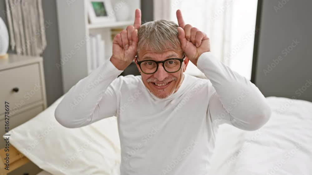 Eccentric middle age, grey-haired man doing a playful bull-horns gesture with fingers over head while wearing pyjamas in bedroom, turning fun bedtime into a funny indoor rodeo. Eccentric middle age, grey-haired man doing a playful bull-horns gesture with fingers over head while wearing pyjamas in bedroom, turning fun bedtime into a funny indoor rodeo.