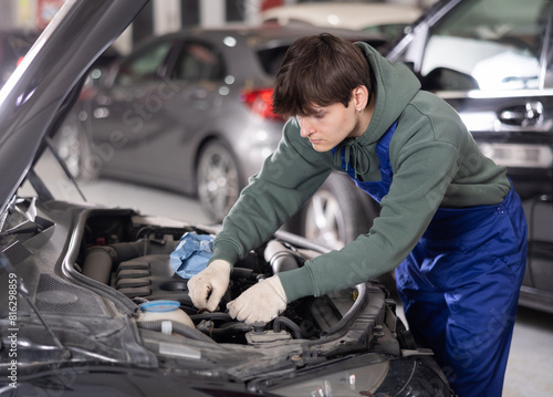 Young male auto mechanic in blue overalls and gloves focused on adjusting parts and tightening bolts under hood of vehicle in repair shop, with blurred background of other cars