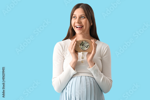 Young pregnant woman with piggy bank on blue background. Maternal Benefit concept