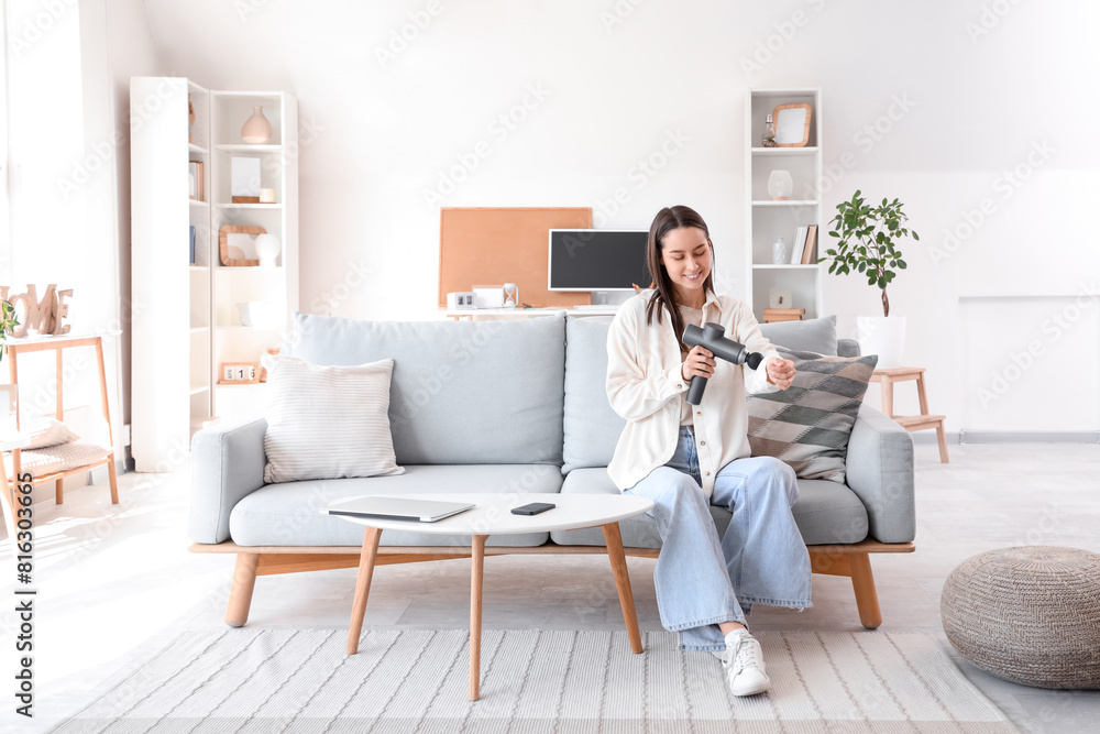 Young woman massaging her arm with percussive massager on sofa at home Young woman massaging her arm with percussive massager on sofa at home