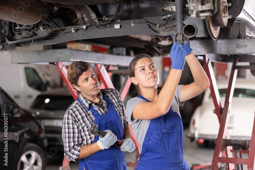 Two focused professional mechanics, middle aged man and young woman, wearing blue overalls, repairing undercarriage of car on lift in auto repair shop.. photo