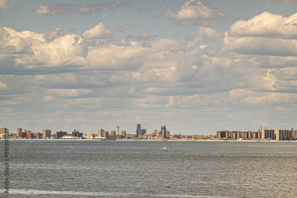 Fototapeta premium New York, NY, USA - August 1, 2023: Maimonides Park on Coney Island beach in wide landscape under blue cloudscape. Gray ocean water up front. Brown buildings belt