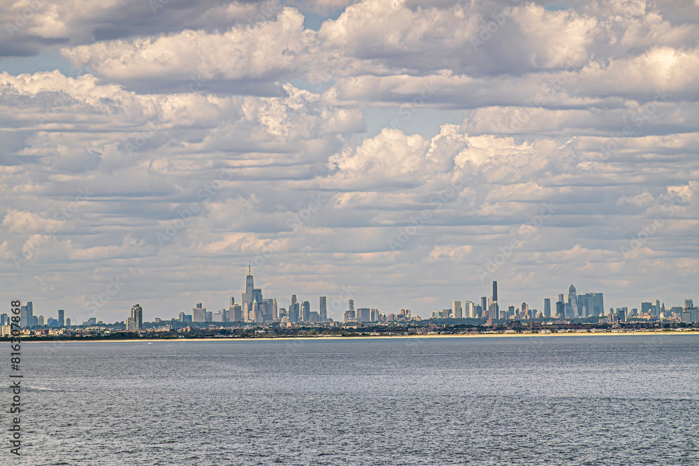 Fototapeta premium New York, NY, USA - August 1, 2023: City skyline off ocean under blue cloudscape. One World Trade center is tallest. Green roof is Federal Hall. Bel Harbor sandy beach line