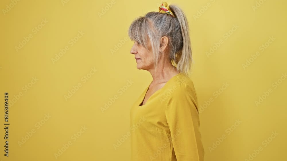 Confident middle-aged woman flashes a natural smile, her grey hair in a ponytail. profile view against a yellow, isolated background. Confident middle-aged woman flashes a natural smile, her grey hair in a ponytail. profile view against a yellow, isolated background.