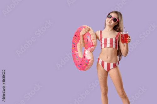 Cute little happy girl in swimsuit with inflatable ring and glass of lemonade on purple background