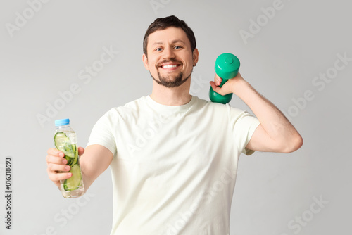 Sporty young man with bottle of cucumber water and dumbbell on light background