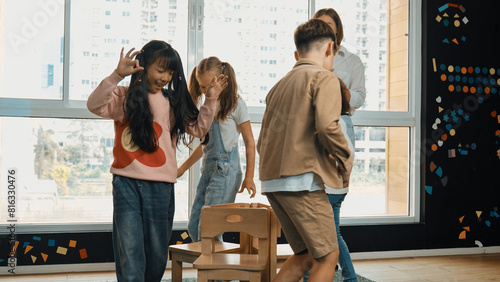 Diverse children playing musical chairs games while young smart beautiful teacher turn off music. Multicultural students running and sitting on chair in creative activity. Music lesson. Erudition. photo