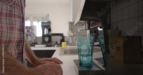 A man is pouring water from a modern filtration device into a clear glass, emphasizing a healthy lifestyle choice in his well-lit kitchen. photo