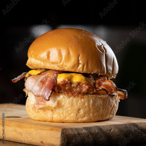 A big hamburger with brioche bread, meat, cheddar cheese, barbecue sauce, and slices of bacon on a wooden table