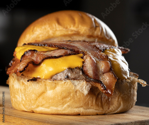 An opened big hamburger with brioche bread, meat, cheddar cheese, barbecue sauce, and slices of bacon on a wooden table