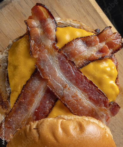 A topdown opened big hamburger with brioche bread, meat, cheddar cheese, barbecue sauce, and slices of bacon on a wooden table