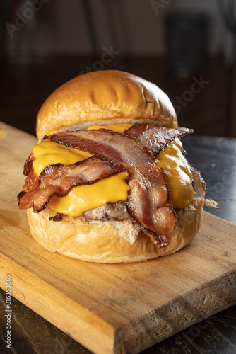 An opened big hamburger with brioche bread, meat, cheddar cheese, barbecue sauce, and slices of bacon on a wooden table