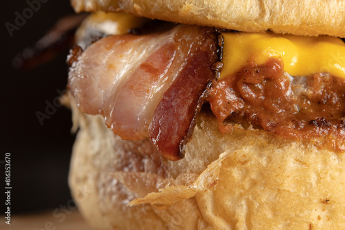 A closeup of a slice of bacon on a hamburger with brioche bread, meat, cheddar cheese, barbecue sauce, and slices of bacon on a wooden table