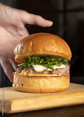 A hand reaching a big hamburger with brioche bread, meat, cheese, and crispy cabbage in a wooden table