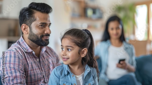 Close up of Indian father sitting in the living room, little daughter standing and talking to him at home, mother blurred in the background holding phone.