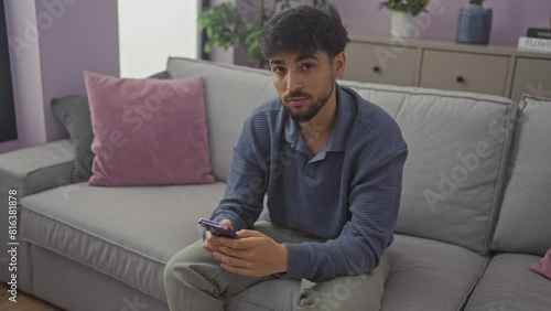 Young arab man, confident and self-assured, immersed in deep thought while sitting on a sofa at home, his smartphone in hand and expression sober photo